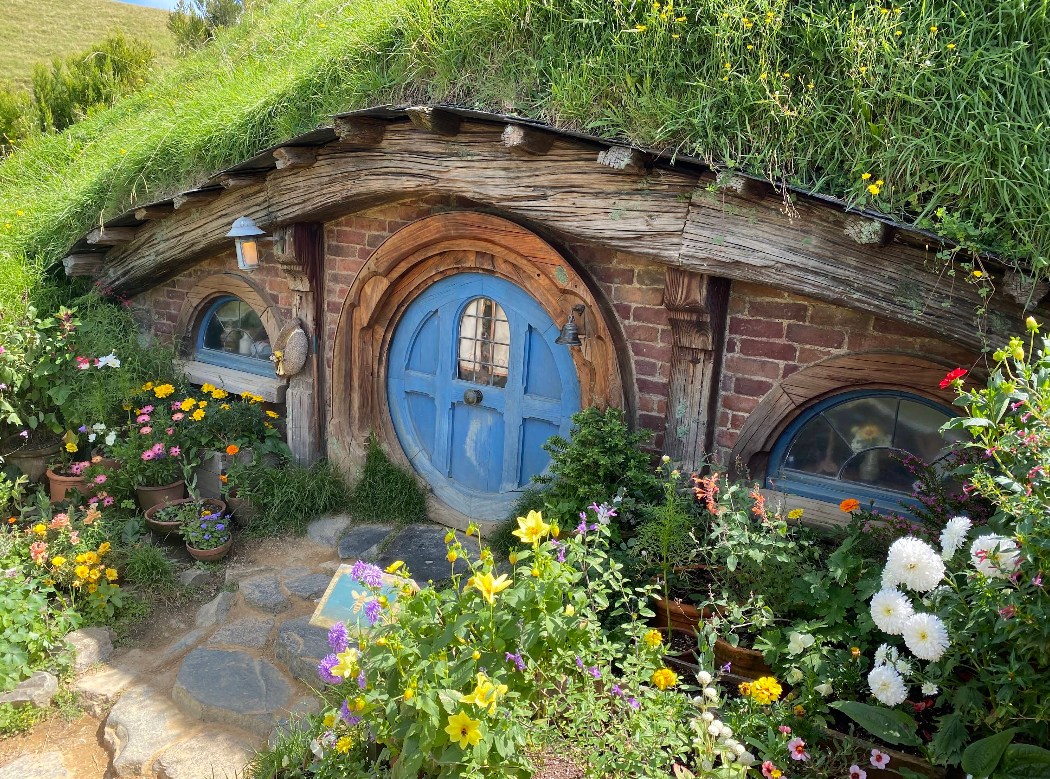 Round blue door of a hobbit-hole home set into a grassy hillside, surrounded by flowers at Hobbiton Movie Set.