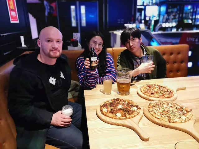 Isaac and friends sharing craft beer and stone-baked pizza at a lively Hamilton dining table.